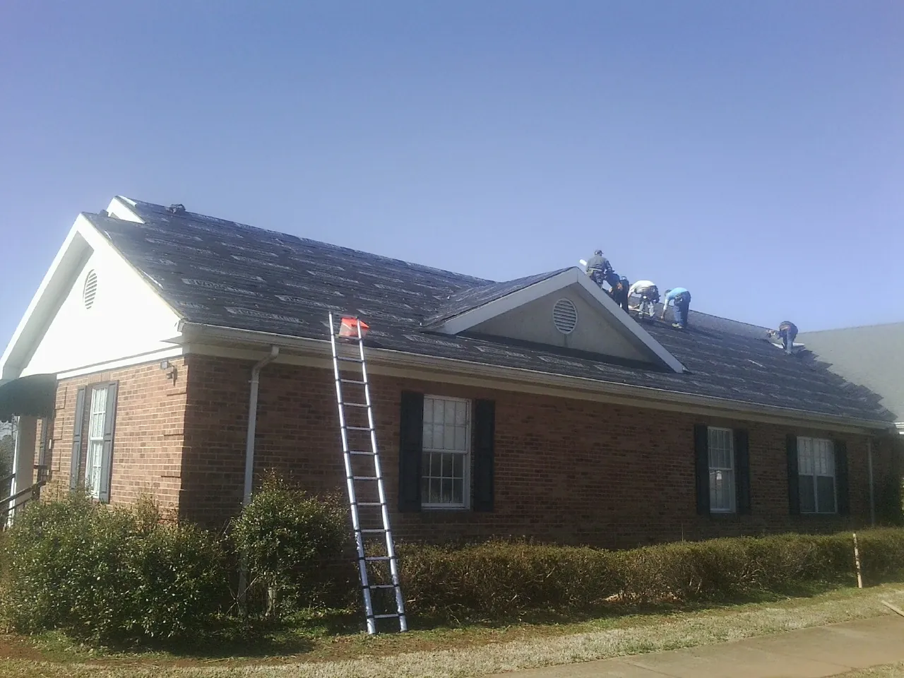Crew tearing shingles off a brick ranch roof with a ladder set — Metal Roof Repair work by our crew in Fort Dodge