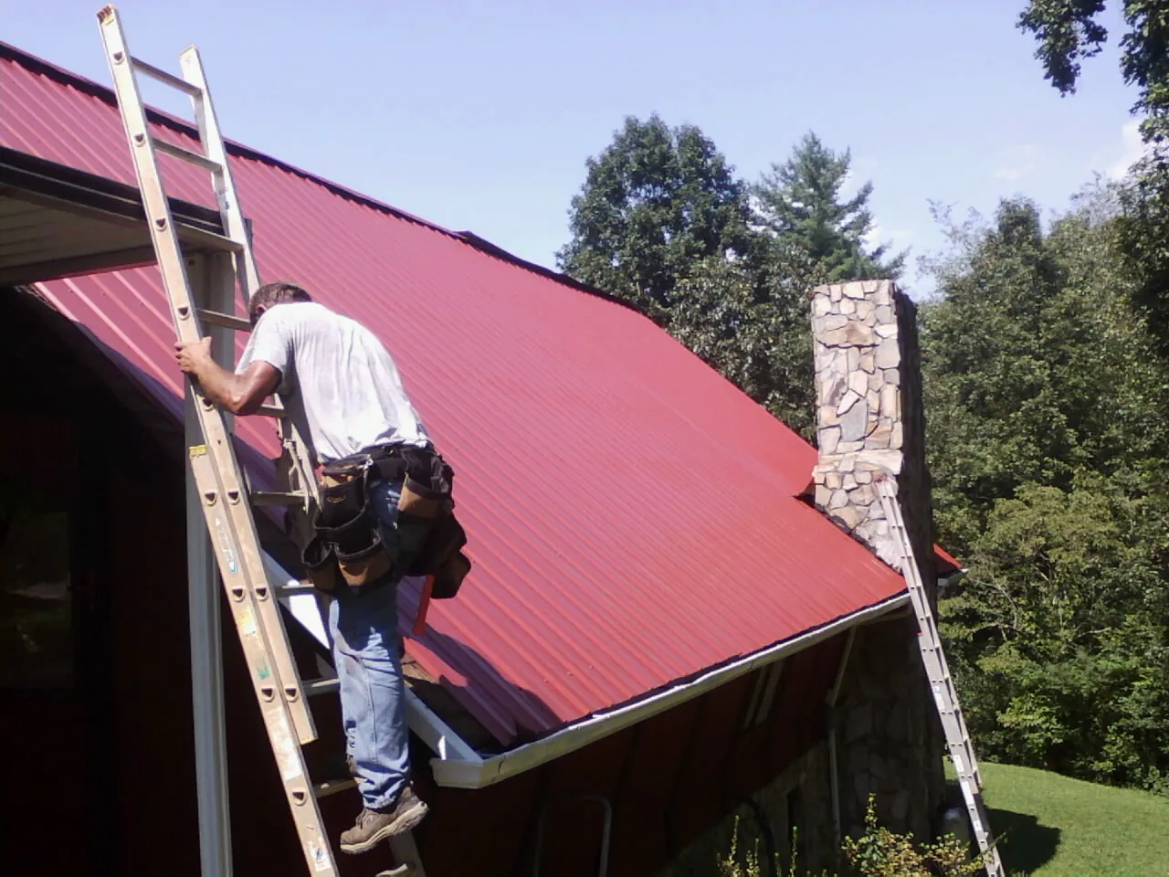 Roofer on a ladder beside a red metal roof and stone chimney — Soffit & Fascia Repair by Roofing Brothers in Fort Dodge